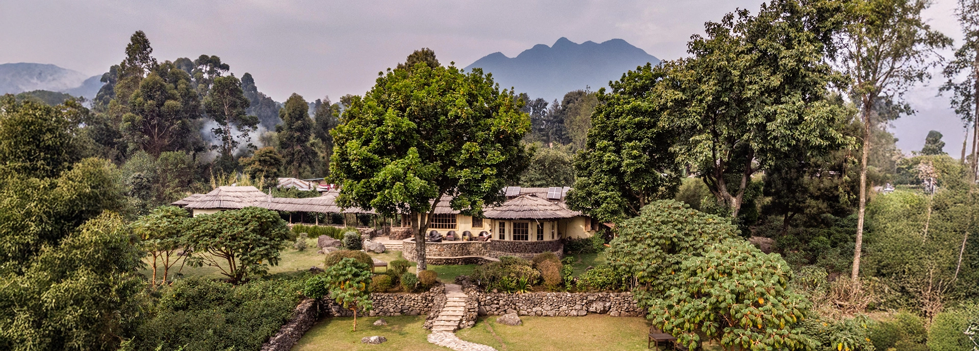 Aerial view of Mount Gahinga Lodge surrounded by lush gardens and forest with the Virunga volcanoes in the background.