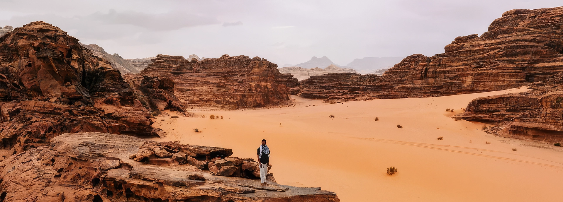 A lone traveller walks across the vast ochre sands and layered rock formations of Wadi Rum beneath a wide desert sky.