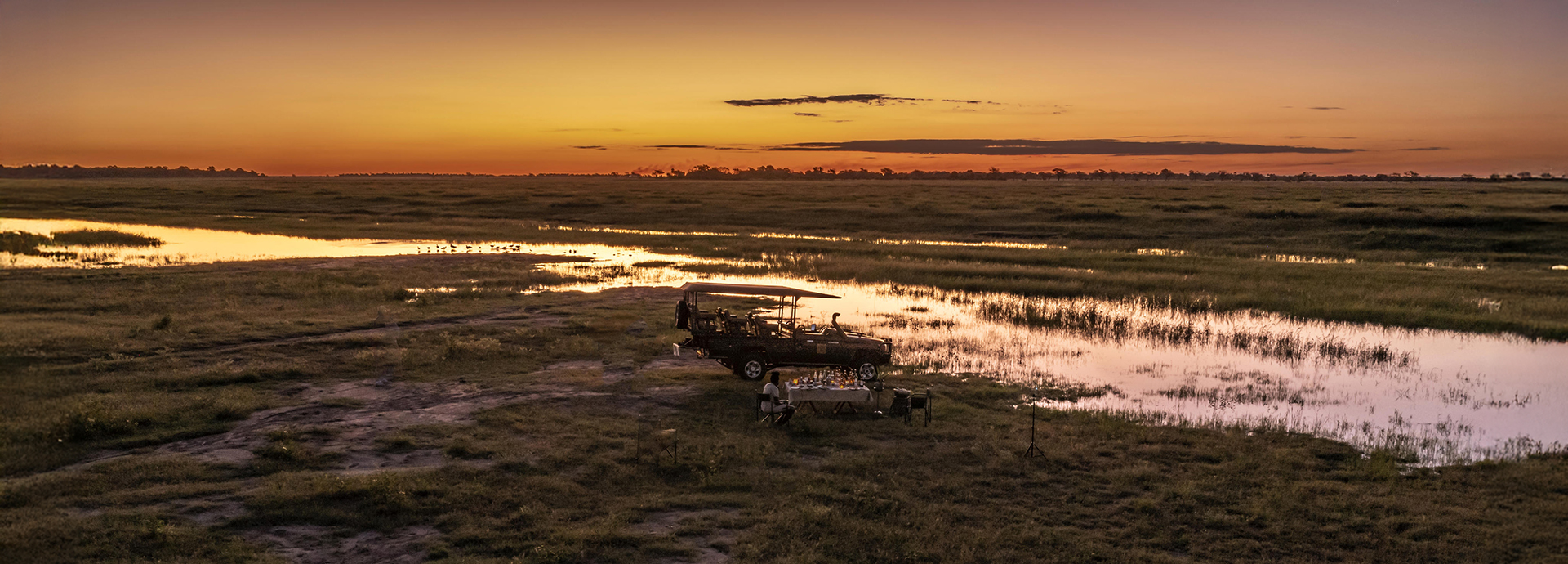 A serene sunset over the Chobe floodplains with a safari vehicle and dining setup for a scenic sundowner experience.