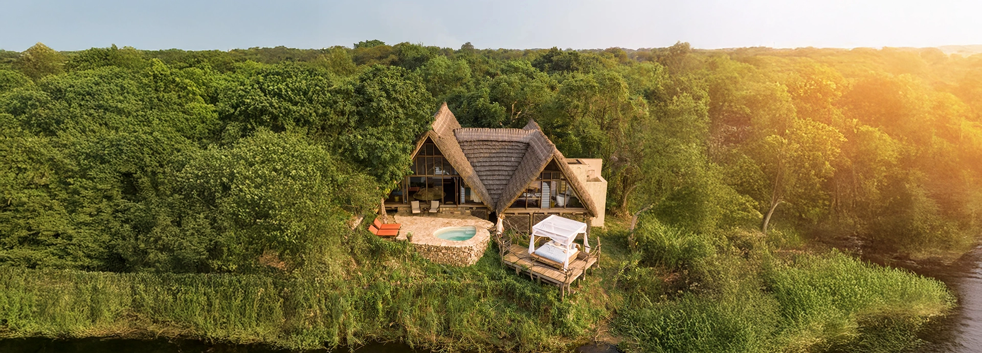 An aerial view of a secluded thatched safari lodge set deep within riverine forest, overlooking the calm waters of the Victoria Nile at golden hour.