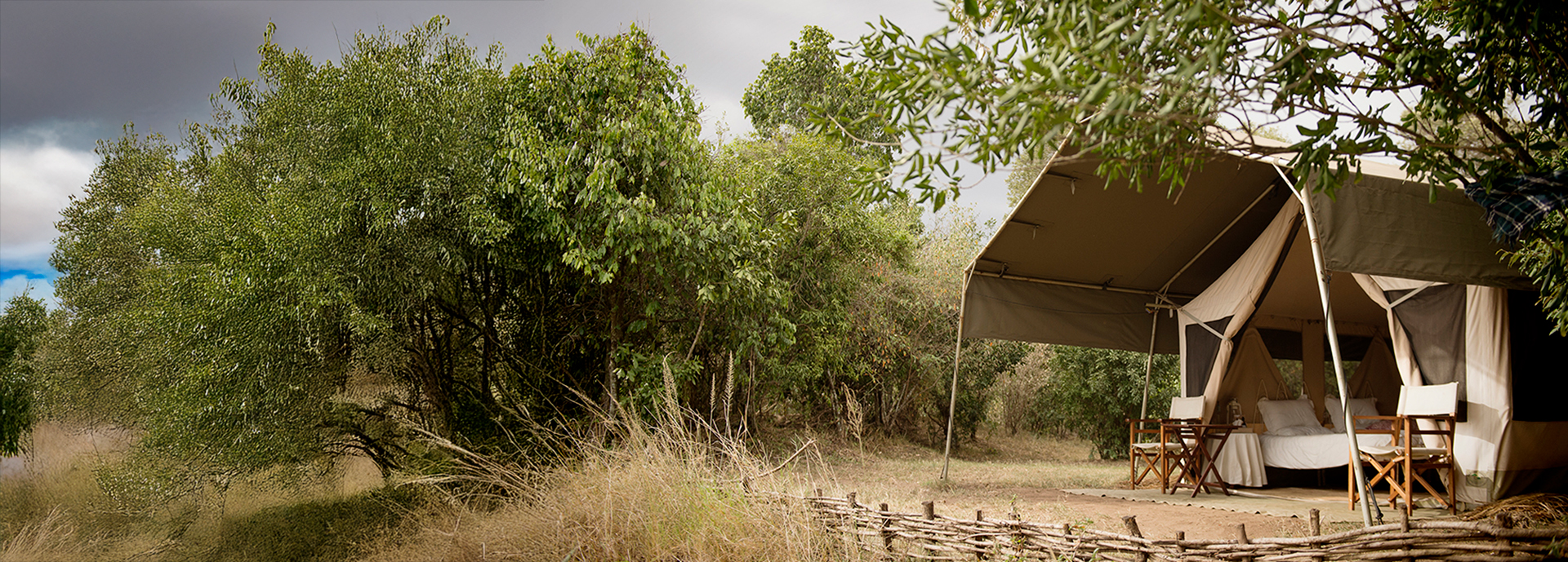 A canvas safari tent with twin beds and wooden chairs set under leafy trees at Nkorombo Mobile.
