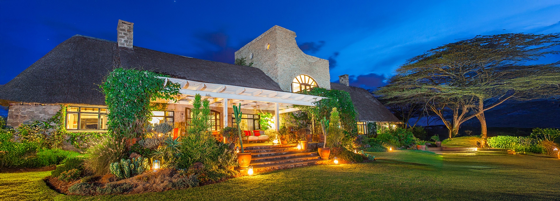 Ololo Safari Lodge illuminated at dusk, its ivy-clad stone walls and veranda glowing beneath a deep blue sky surrounded by lush gardens.