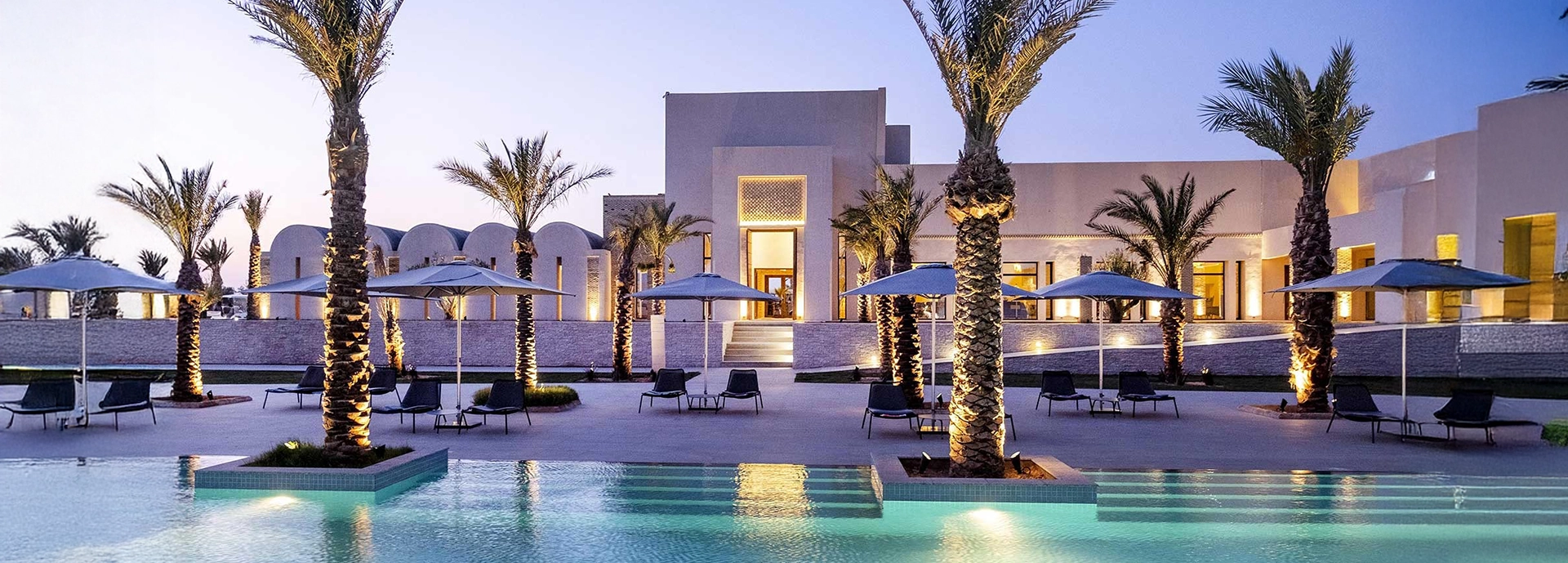 Twilight view of The Residence Douz main pool framed by palm trees and softly lit architecture.