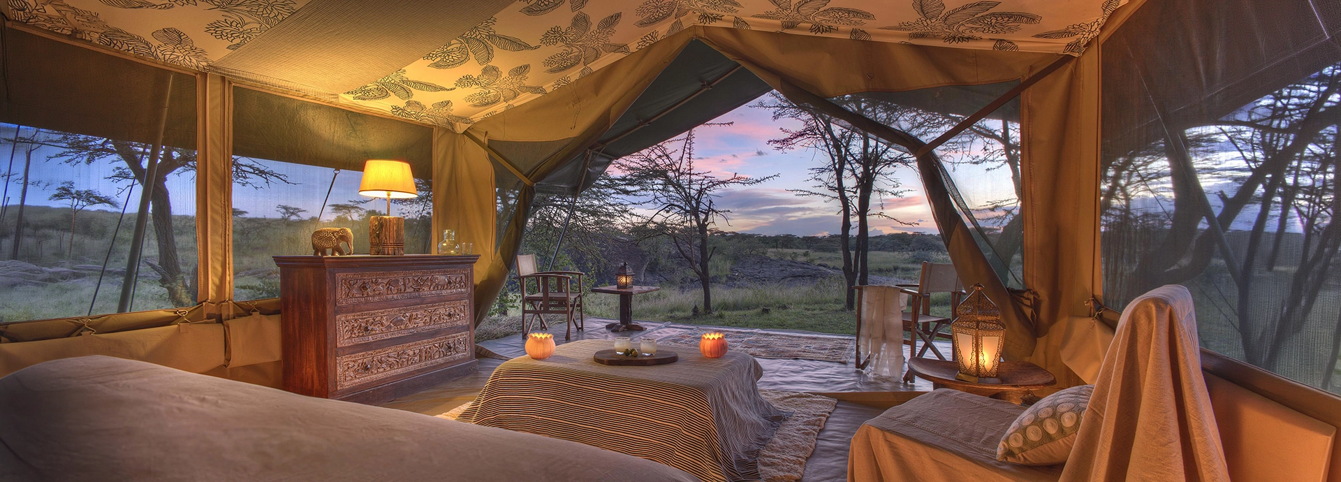 A warmly lit luxury safari tent interior at dusk, with carved wooden furniture, candlelight and open canvas sides framing a rose-coloured Mara sky.