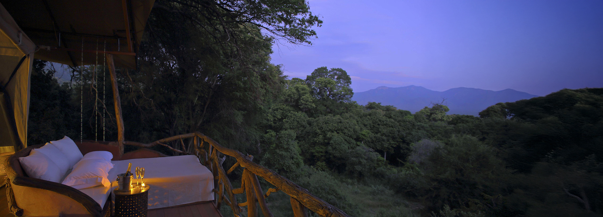 A twilight view from a treehouse deck with a cosy daybed, overlooking the lush forest and distant mountains of the Mathews Range.