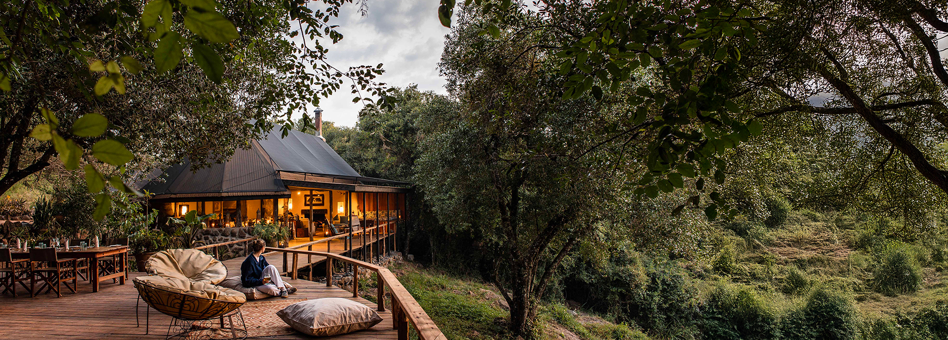 A woman relaxes on a wooden deck outside the warmly lit main tent at Serian ‘The Original’, surrounded by forest.