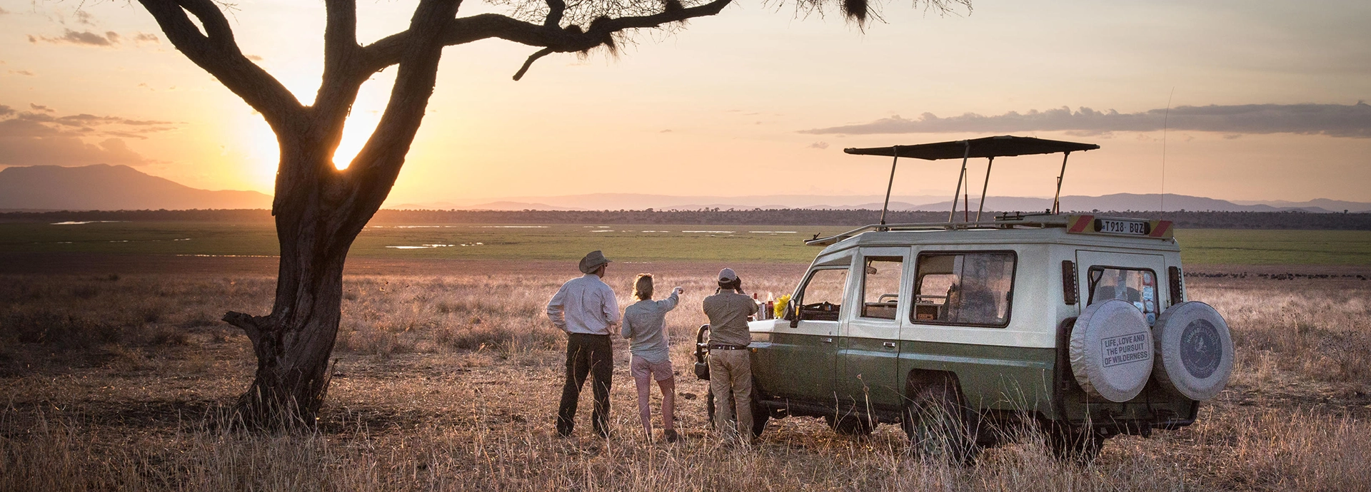 Travellers enjoy sundowners beside a safari vehicle as the sun sinks behind distant mountains and wide open Tarangire plains.