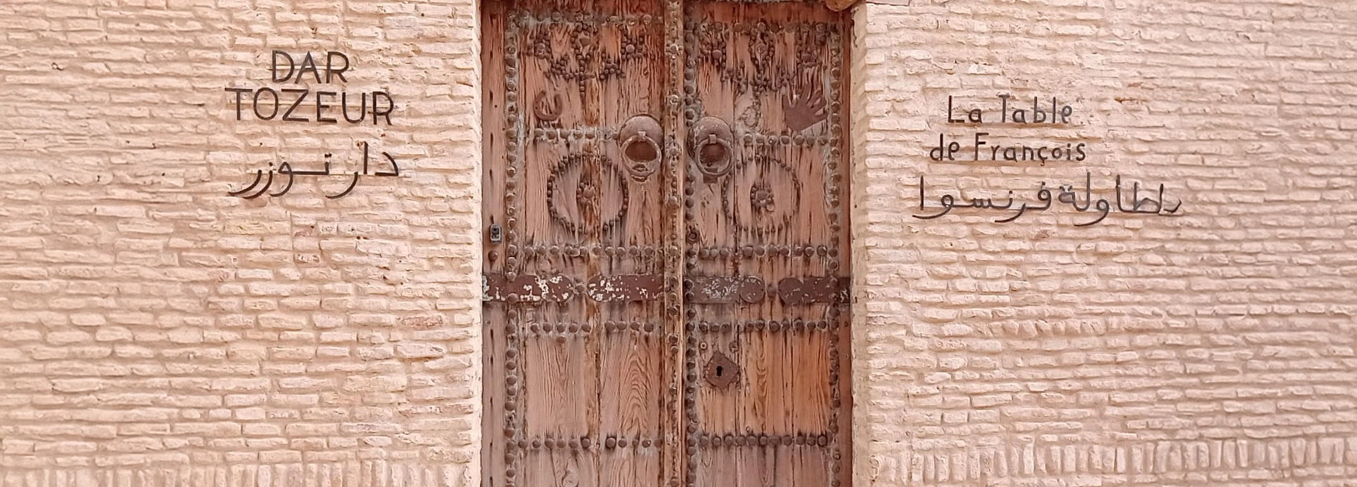 Intricately carved wooden entrance door to Dar Tozeur framed by sand-coloured brick walls.