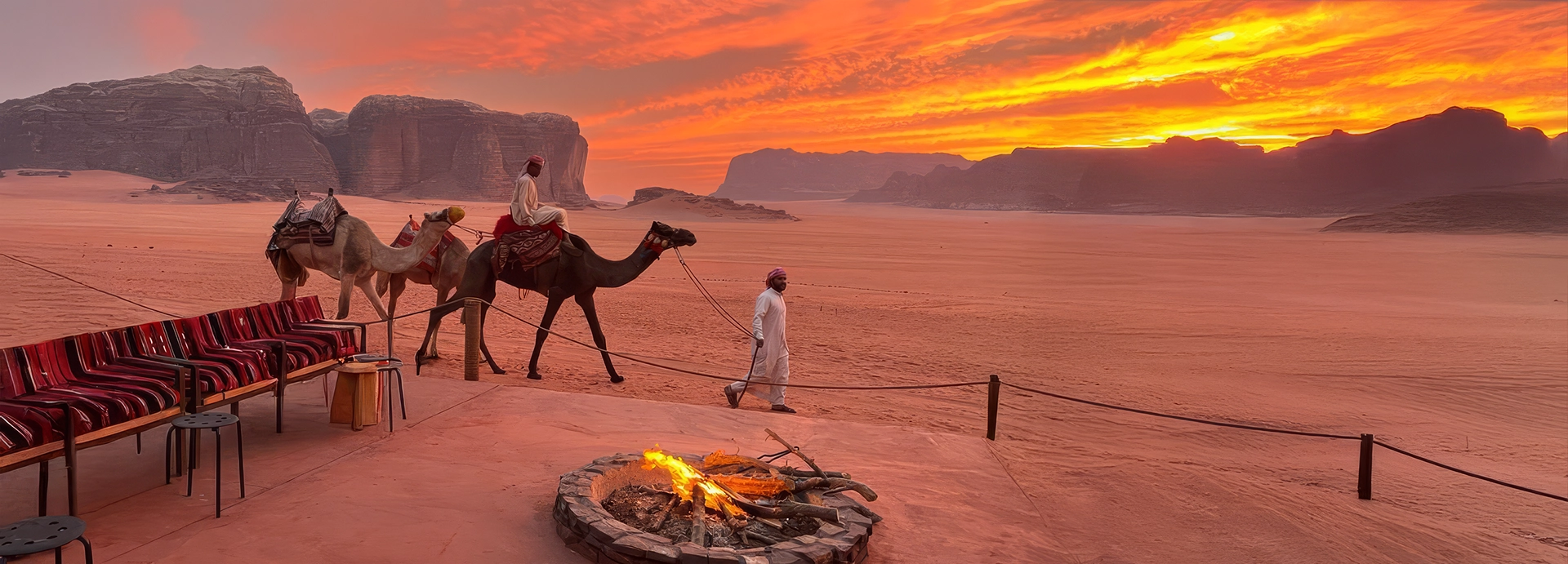 Camels pass the camp fire pit at sunset as the sky ignites with colour across the open desert landscape of Wadi Rum.