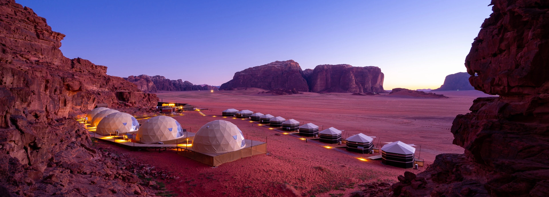 The camp stretches across the desert floor at dusk, with softly lit tents leading towards distant sandstone mountains.