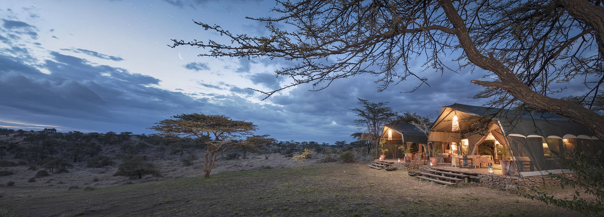 A warmly lit canvas tent glows against a dramatic dusk sky at Hemingways River Camp Mara, surrounded by acacia trees and the darkening Mara bush.