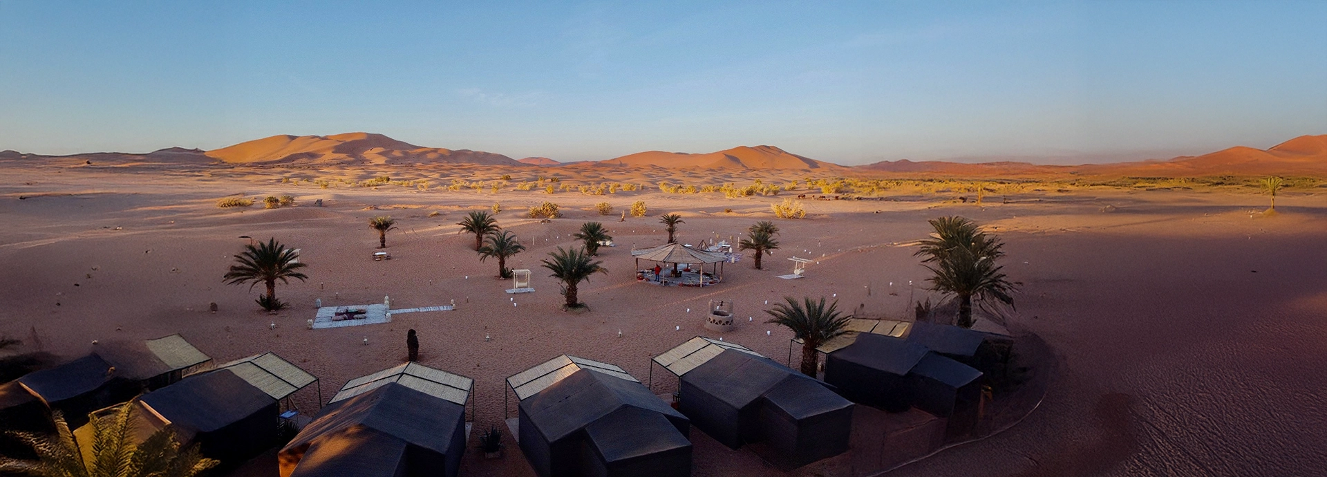 Wide view of Riad Madu Luxury Desert Camp with palm trees and surrounding golden Sahara dunes.