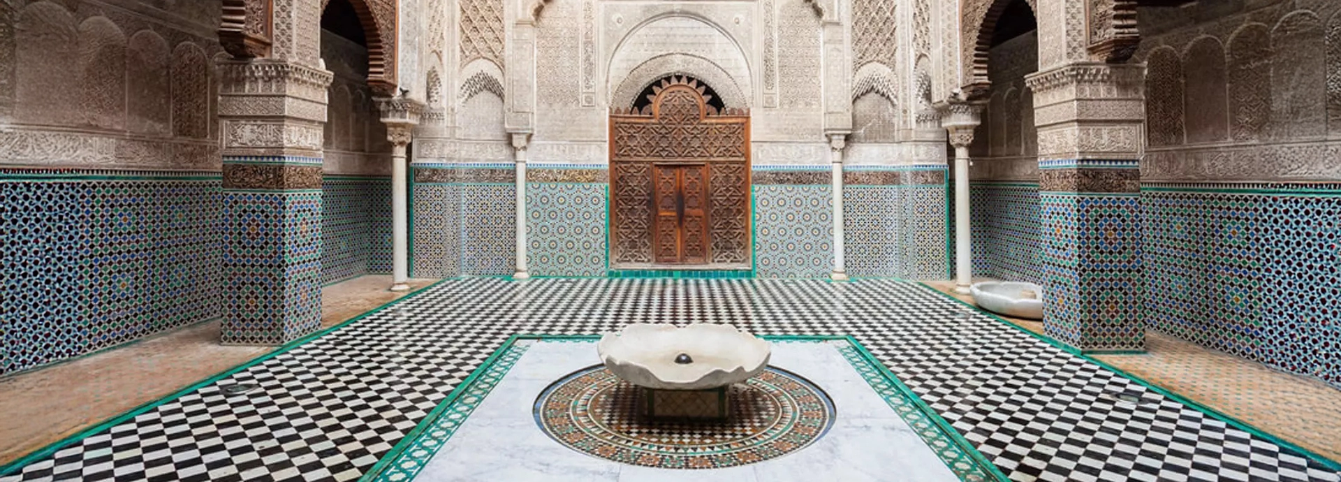 Intricately tiled courtyard of Al Attarine Madrasa in Fès, featuring detailed arches and a central marble fountain.