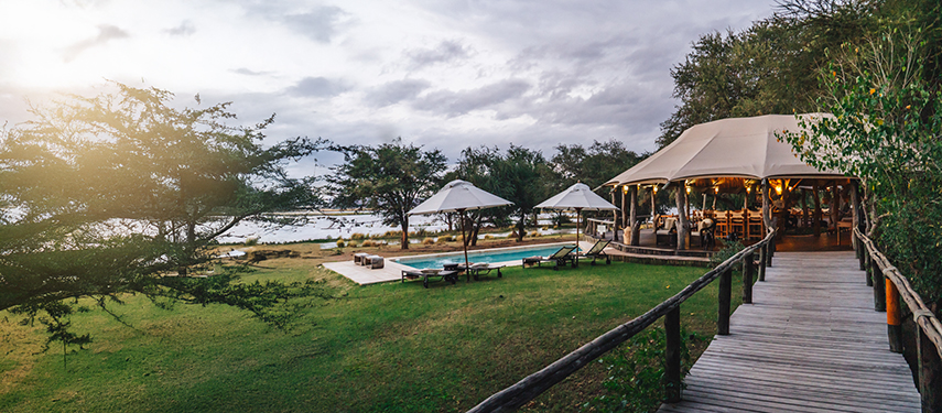 Dulini Anabezi camp's main lounge and pool area overlook the river, lit warmly as the sun sets.