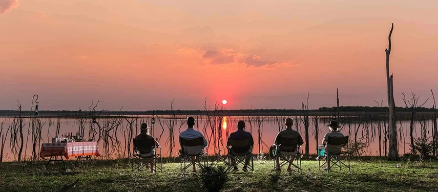 Guests seated in safari chairs watching the sun set over Lake Kariba, with flooded trees rising from the water’s surface.