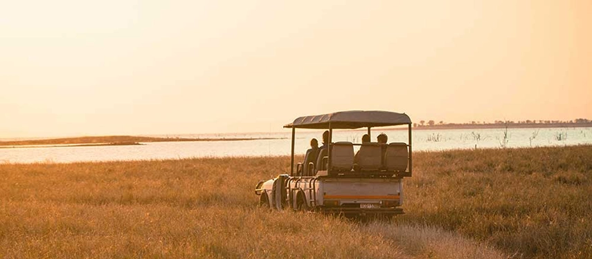 Open-sided safari vehicle carrying guests across golden grasslands beside Lake Kariba during late afternoon.