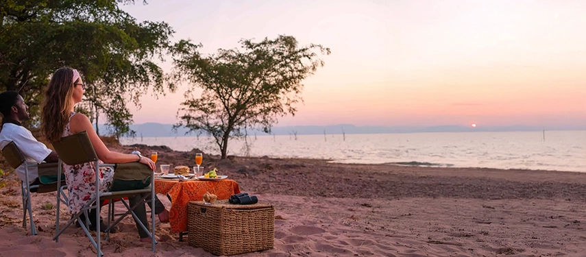 Couple enjoying drinks on the sandy lakeshore at sunset, with a simple table set and Lake Kariba stretching beyond.