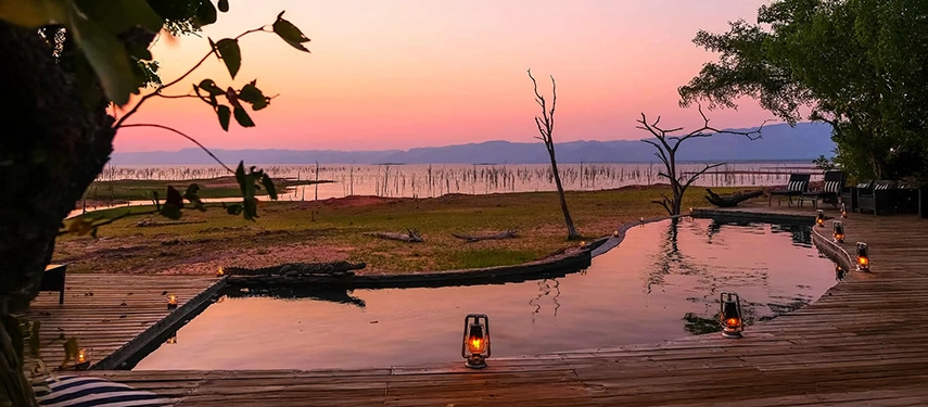 Infinity pool at Changa Safari Camp overlooking Lake Kariba at sunset, with lanterns lining the timber deck and water stretching into the horizon.
