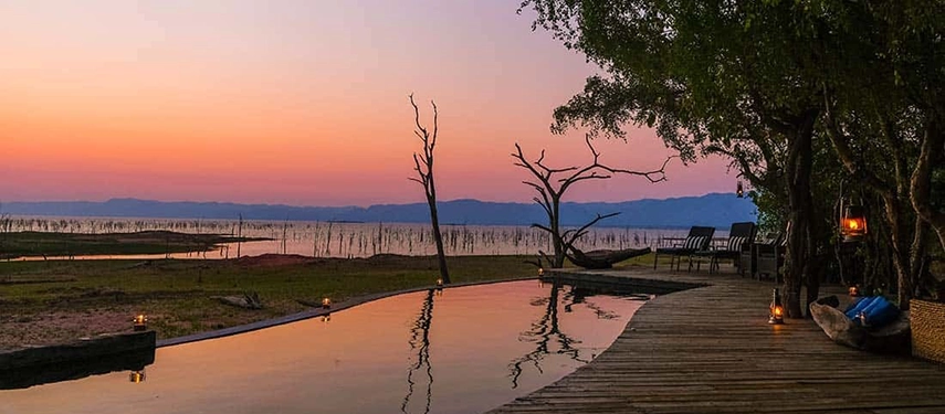 Infinity pool at Changa Safari Camp overlooking Lake Kariba at dusk, framed by lantern-lit decking and silhouetted trees.