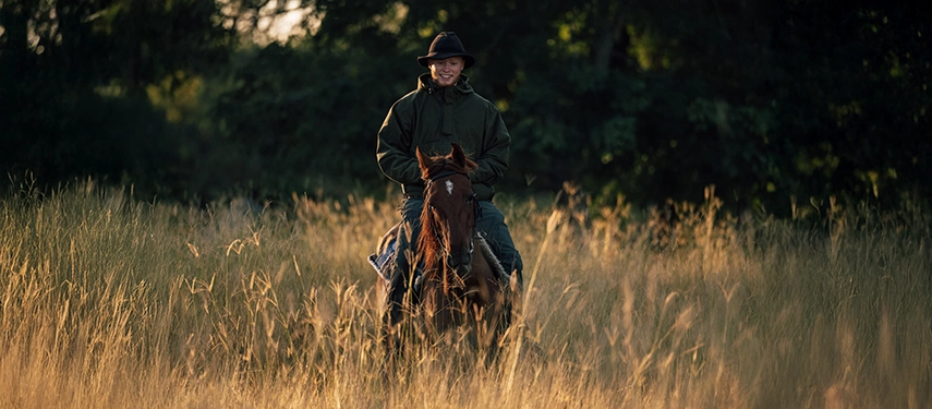 Horse rider smiling through tall golden grass in the soft morning light of western Zambia.