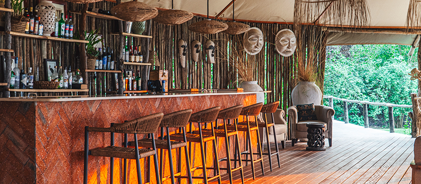The bar at Dulini Anabezi Camp features woven stools, tribal masks, and rustic wooden decor under canvas.