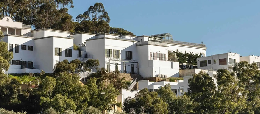 View of Dorp Hotel’s hillside buildings surrounded by greenery, with whitewashed walls and a glass conservatory.