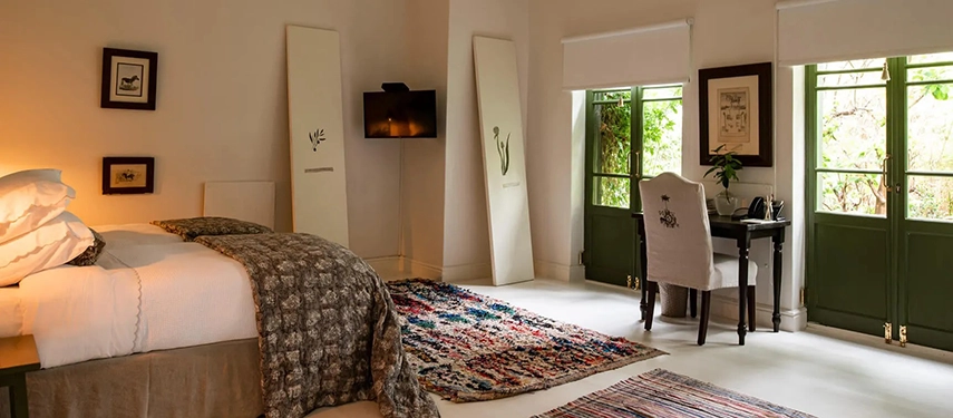 Cosy Dorp Hotel bedroom with floral upholstery, vintage rugs, and French doors opening to sweeping city views.
