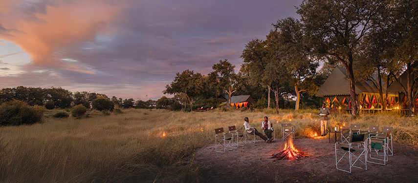 Evening view of the firepit area with guests seated, lanterns glowing, and the camp tents softly lit.