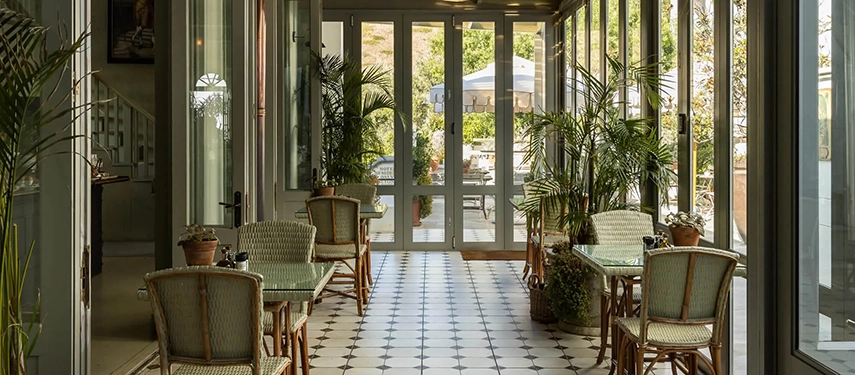 Sunlit conservatory dining area at Dorp Hotel with black-and-white tiled floors, rattan chairs, and potted palms.