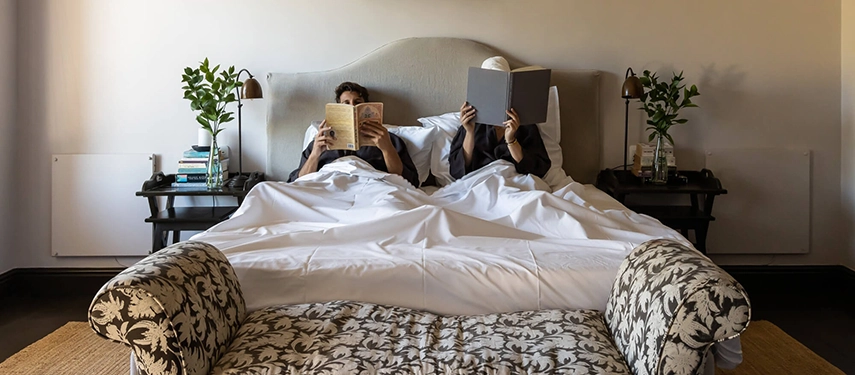 Couple reading in bed at Dorp Hotel, surrounded by natural light, soft linens, and understated, elegant furnishings.
