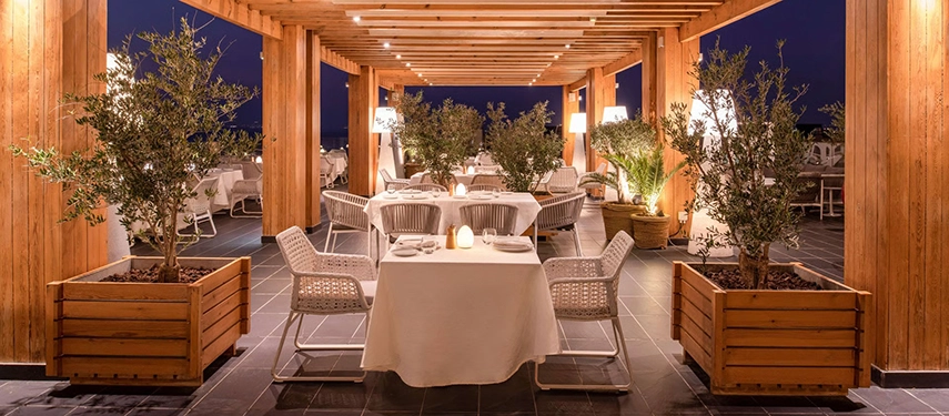 An elegant al fresco dining terrace framed by timber pergolas, soft lighting, and potted olive trees overlooking the Dead Sea at night.