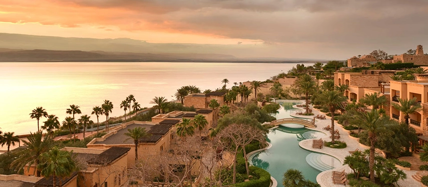 A panoramic view across the resort’s winding pools and palm-lined terraces, descending towards the glowing waters of the Dead Sea at sunset.
