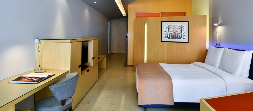 A contemporary guest room with clean lines, pale stone floors, and a writing desk set beside a neatly dressed bed.