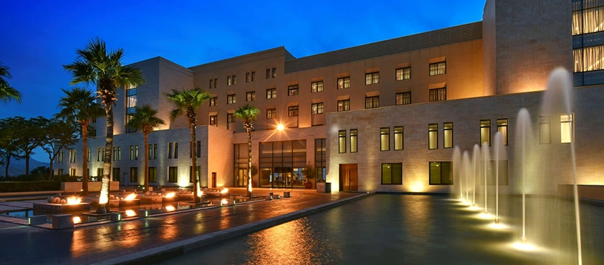 The illuminated façade of Kempinski Hotel Ishtar Dead Sea reflected in still water features, with palm trees and fountains glowing at dusk.