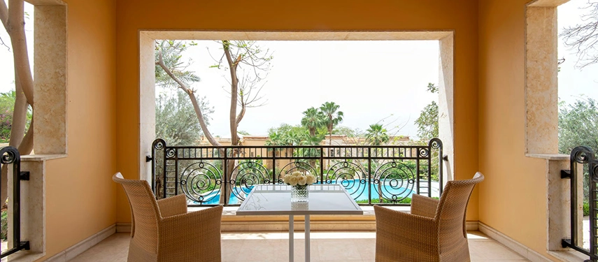 A private balcony seating area framed by warm-toned walls, overlooking landscaped gardens and palm trees with a glimpse of the Dead Sea beyond.