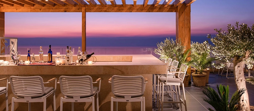 An elegant open-air bar beneath a timber pergola, overlooking the Dead Sea at sunset with soft lighting and relaxed seating.
