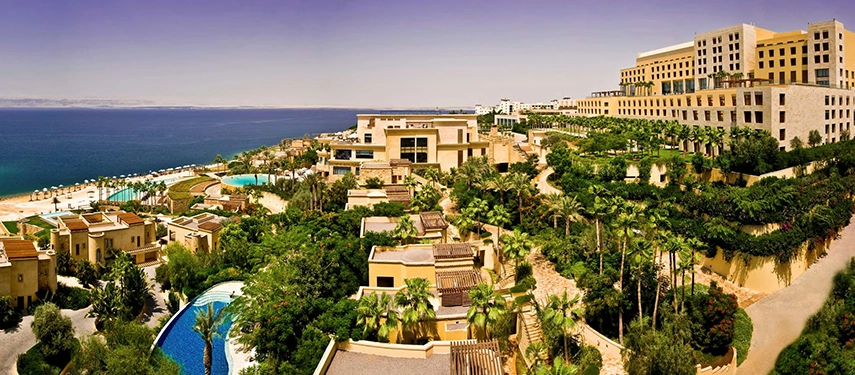 A sweeping aerial view of Kempinski Hotel Ishtar Dead Sea, with terraced buildings, lush gardens, pools, and the Dead Sea stretching into the distance.