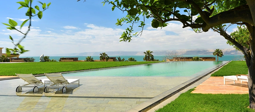 A serene infinity pool set within manicured lawns, opening towards sweeping views of the Dead Sea under a clear blue sky.