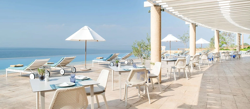 Sun loungers and shaded tables arranged along a bright terrace above the Dead Sea, offering relaxed daytime dining with panoramic views.