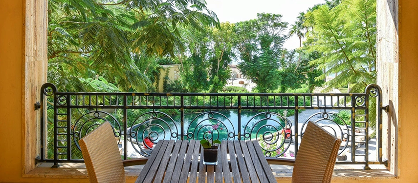 A private balcony dining table overlooking lush gardens and a turquoise pool, framed by ornate iron railings and leafy trees.