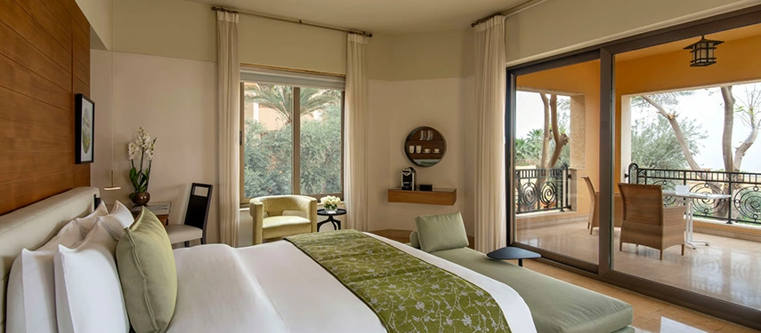 A light-filled guest bedroom with soft green accents, floor-to-ceiling doors, and a private terrace overlooking gardens and palms.