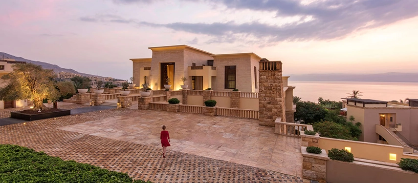 A grand stone courtyard and palace-style residence overlooking the Dead Sea at sunset, softly lit with warm tones and expansive terraces.
