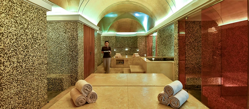 The spa hammam interior with mosaic-tiled walls, vaulted ceilings, stone treatment tables, and neatly rolled towels in a tranquil setting.
