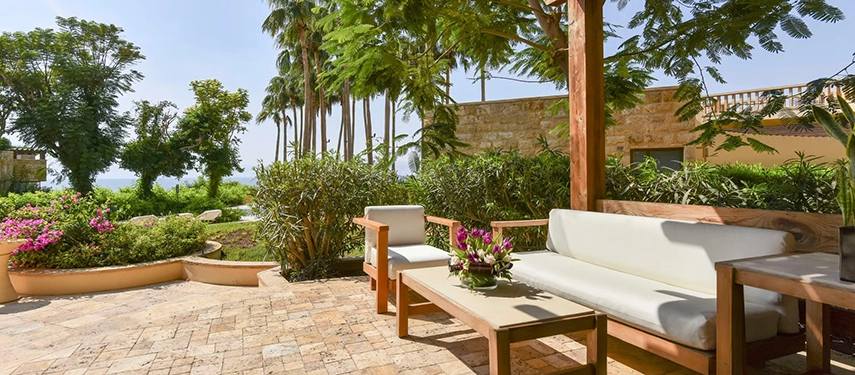 A shaded garden terrace with cushioned outdoor seating, flowering plants, and palm trees creating a peaceful retreat near the Dead Sea.