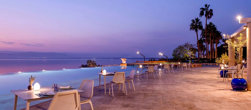 Infinity pool terrace at twilight with softly lit tables and sweeping views across the still waters of the Dead Sea.