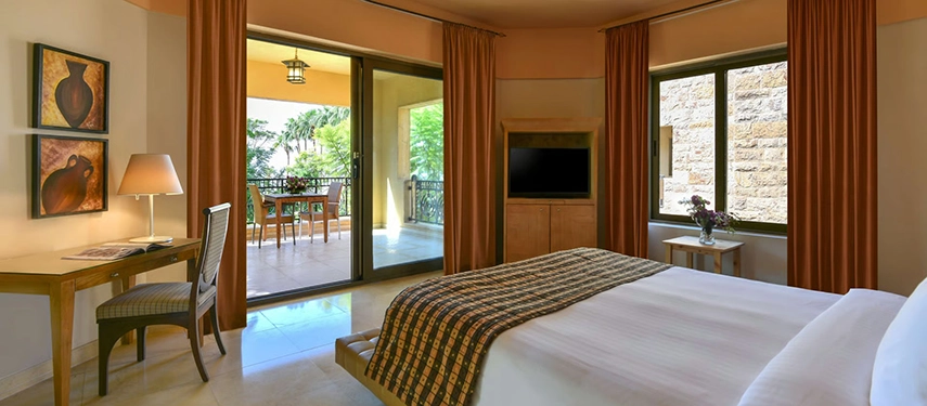 A refined guest bedroom with warm tones, stone accents, and a furnished balcony overlooking landscaped gardens.