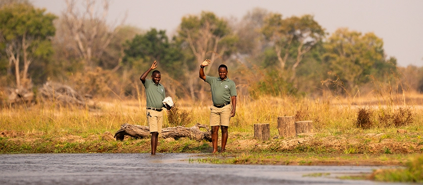 Two camp team members wave from the riverbank, capturing the warm and welcoming spirit of Kutandala Camp.