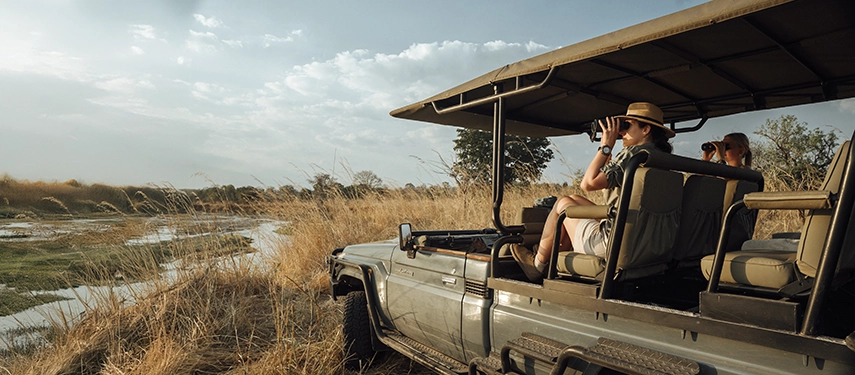 Guests observe the landscape from a safari vehicle parked beside a winding river, binoculars raised in search of game.