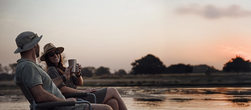 Guests relax with drinks beside a river at sunset, enjoying a quiet moment as the Luangwa landscape softens into evening light.
