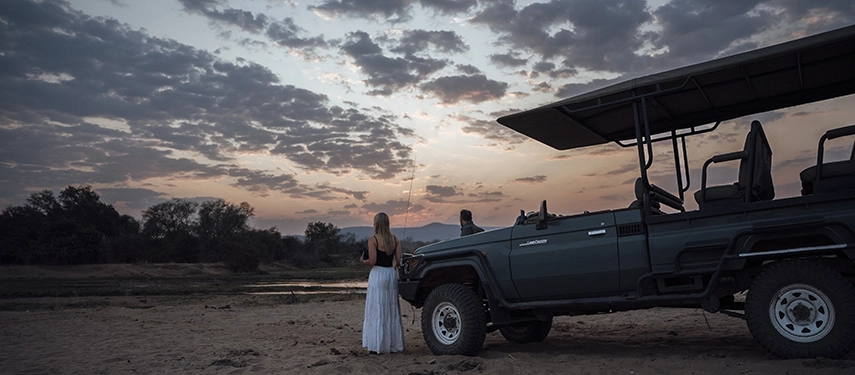 Guests stand beside a safari vehicle at sunset, watching the river and sky change colour at the end of a day in North Luangwa.